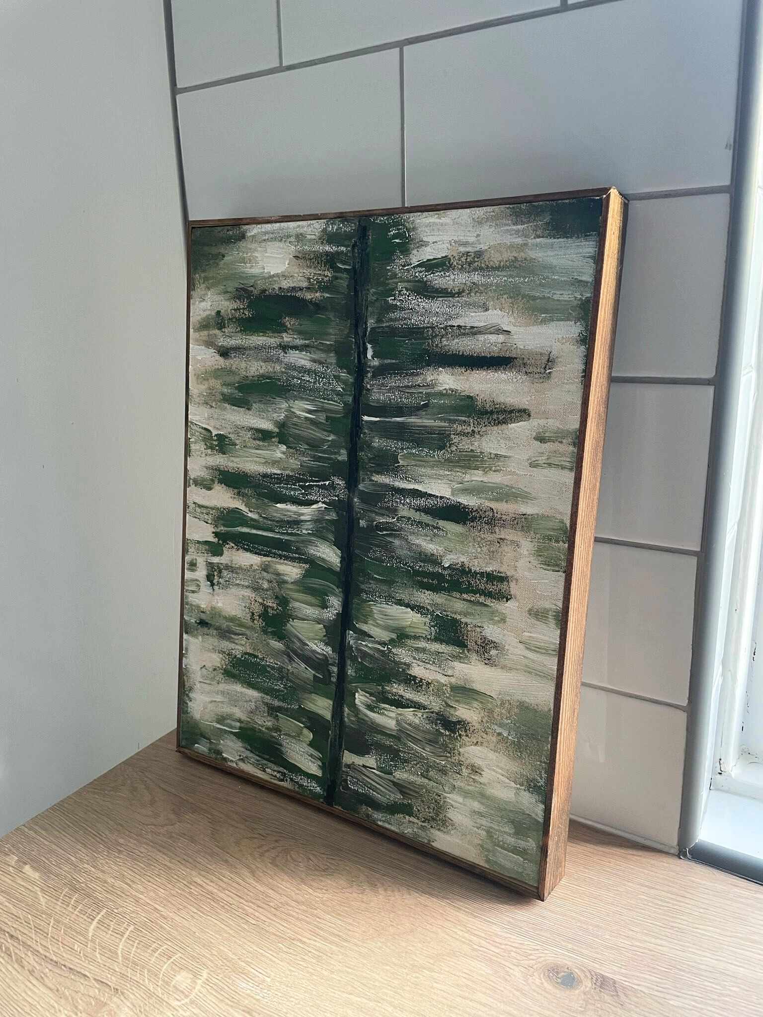 Customer creation made with a Plan framed canvas kit, abstract green and grey brushstroke painting on canvas with an oak edge frame, leaning on a wooden countertop by a window