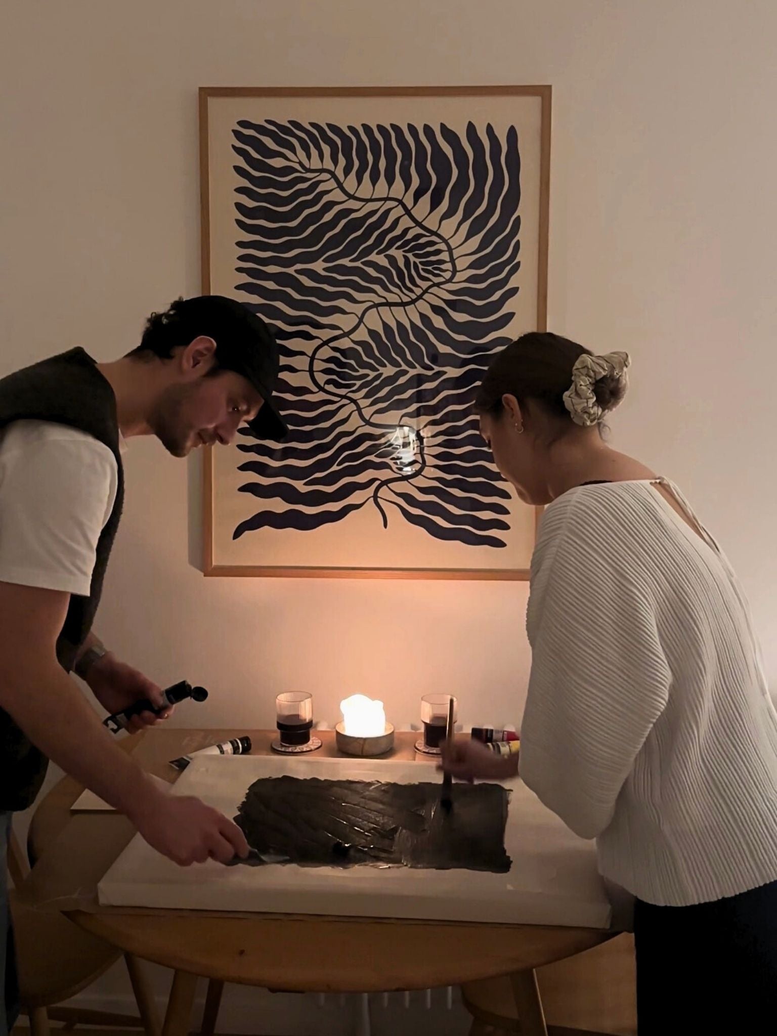 Two people painting together at a dining table using a Plan DIY framed canvas art kit, relaxed evening setup with candles showing a calm, shared creative moment and date night at home