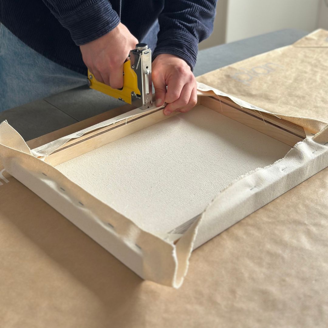 Stretching raw cotton canvas onto a custom wooden frame using a staple gun as part of a DIY framed canvas art kit.