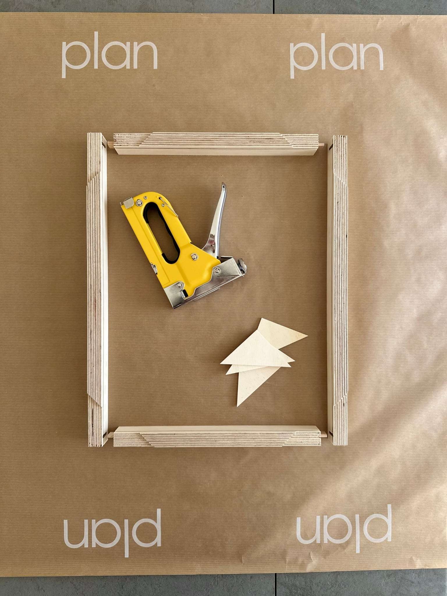 Plan DIY framed canvas art kit inner frame laid out with wooden stretcher bars, corner wedges and staple gun, shown on branded protective paper