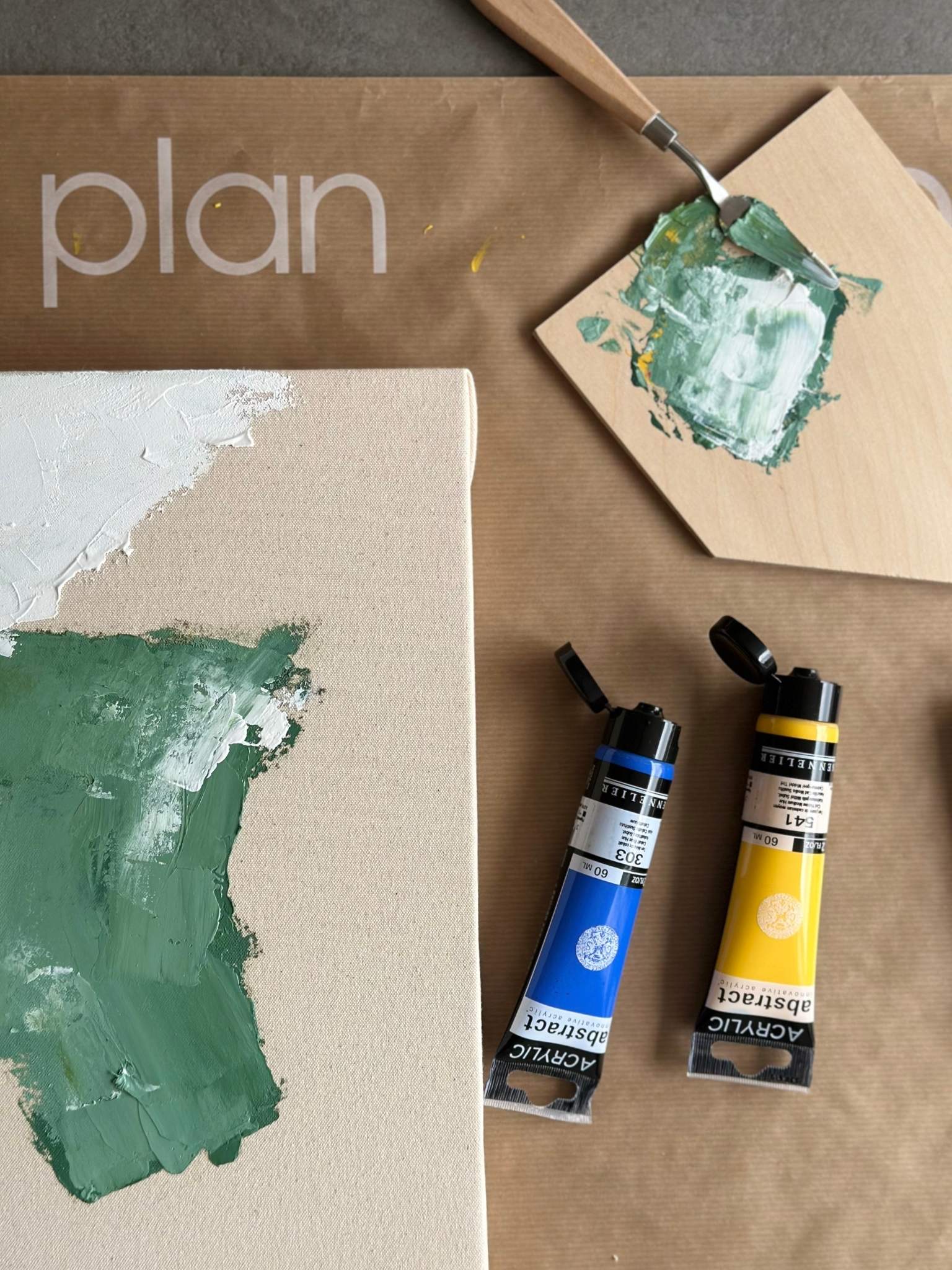 Process shot of abstract canvas painting in progress using the Mix framed canvas art kit, showing green and white acrylic paint applied to raw canvas with palette knife, alongside blue and yellow acrylic paint tubes.