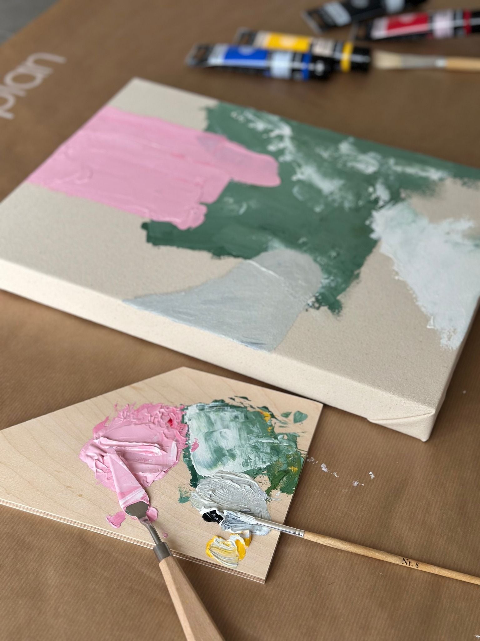 Abstract canvas painting in progress using primary colours, showing mixed green, pink and white acrylic paint on canvas, with paint palette and tools laid out during the creative process.