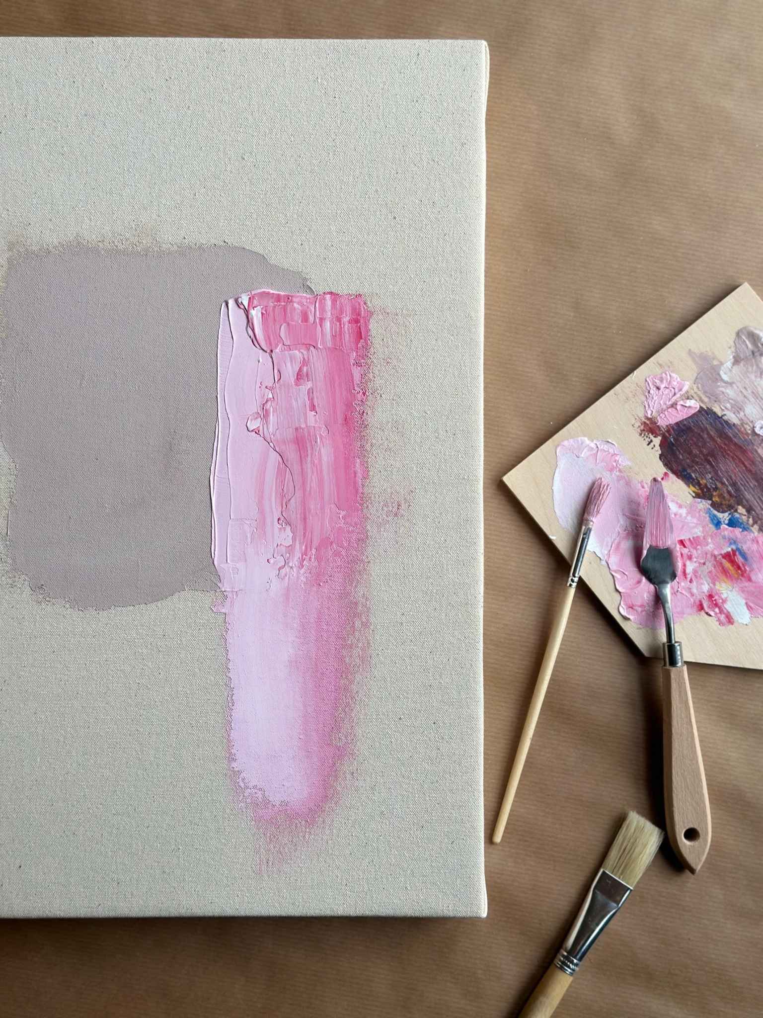 Detail shot of abstract painting process using the Mix canvas art kit, featuring soft grey and pink paint layers on raw canvas with brushes and palette knife nearby.