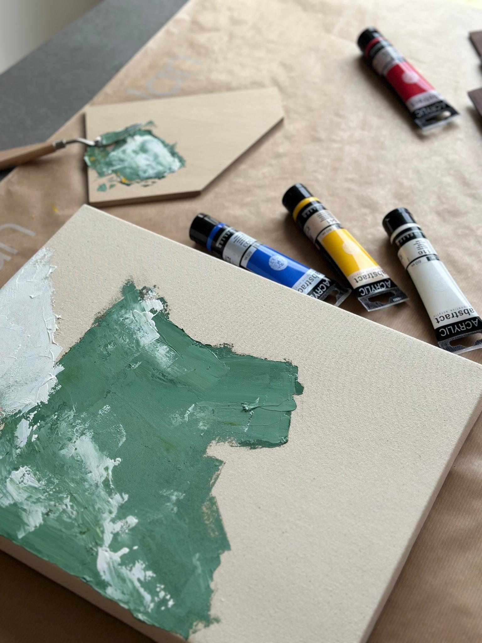 Process shot of a DIY framed canvas art kit using primary colour acrylic paints, showing green paint being mixed and applied to raw canvas with red, blue, yellow and white paint tubes nearby.
