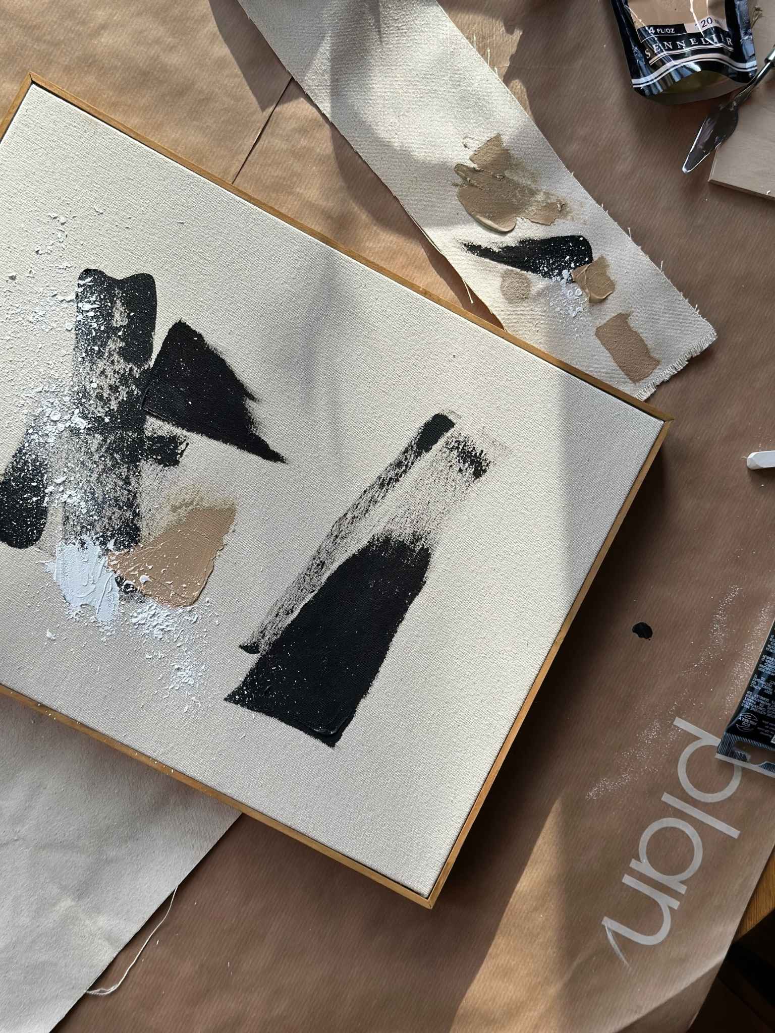 Top-down process shot of Plan DIY art kit abstract painting in progress on cotton canvas, featuring black, white and beige paint marks with palette knife and paint test strip nearby.