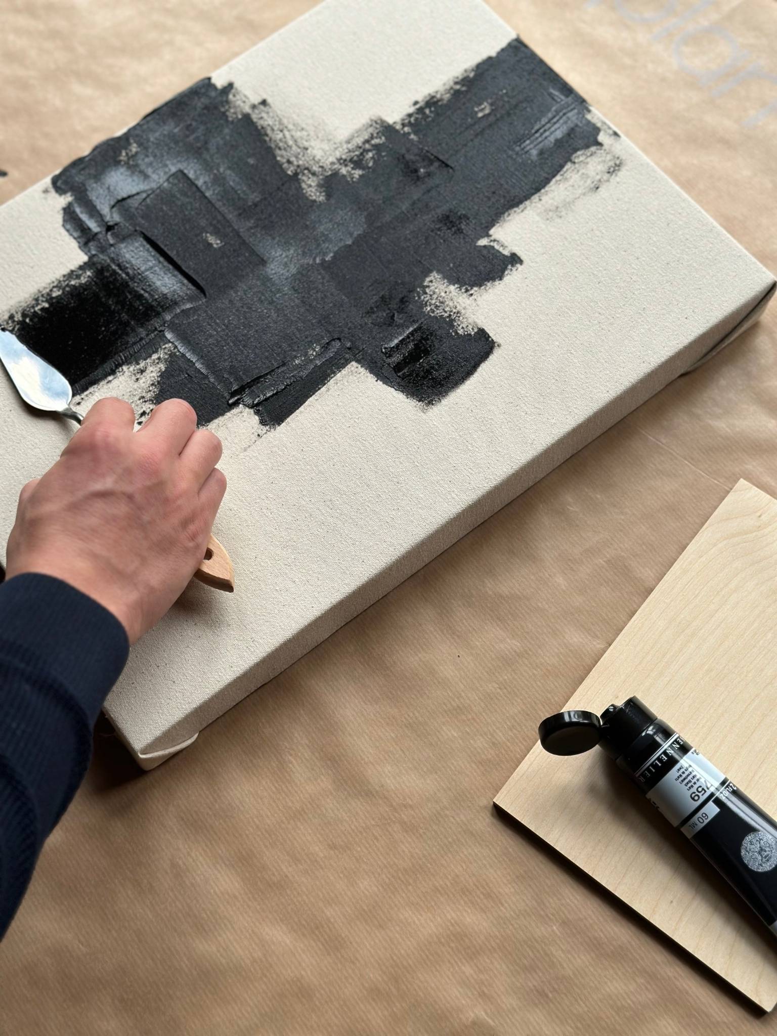 Process shot of hand using a palette knife to apply black acrylic paint onto beige cotton canvas, demonstrating expressive mark-making.