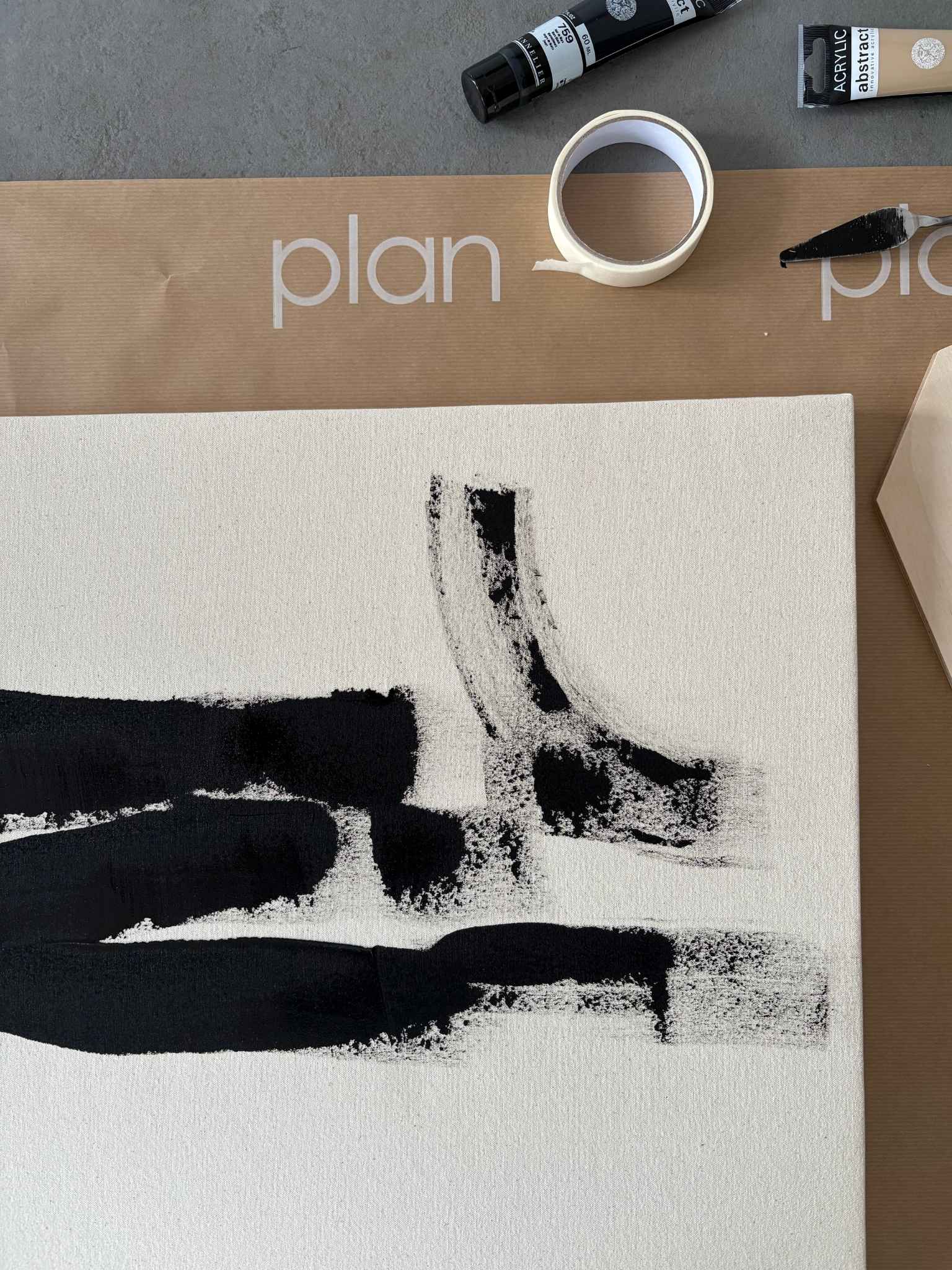 Close-up process shot of monochrome abstract painting in progress, featuring black brushstrokes on beige cotton canvas with masking tape, paint tube and tools visible.