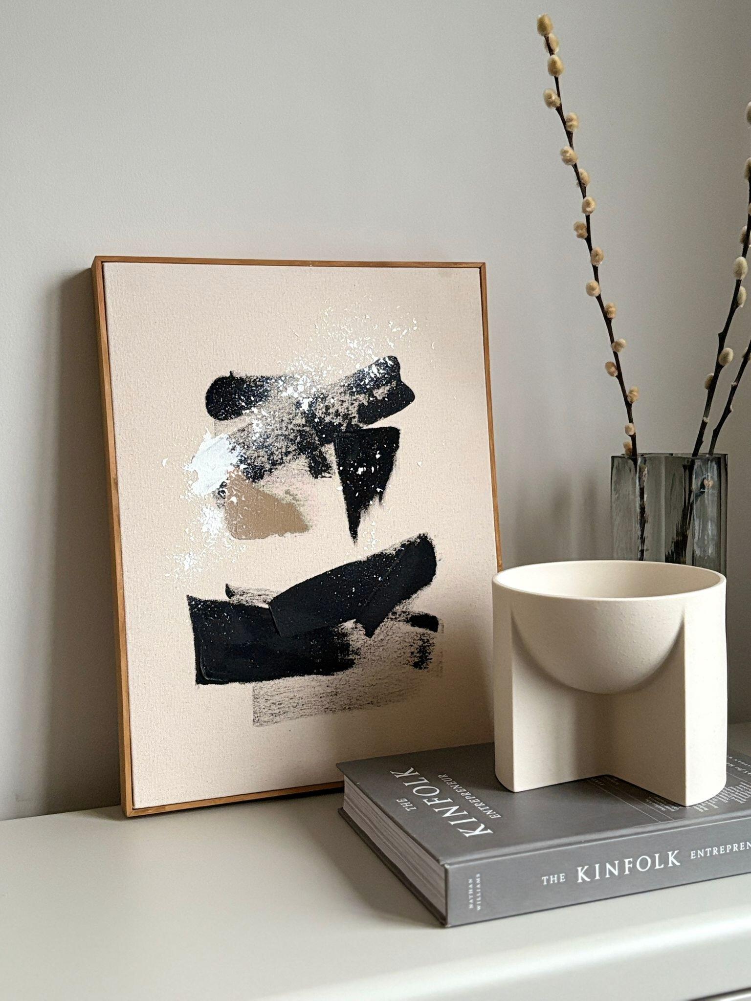 Abstract monochrome framed canvas artwork leaning on a shelf, featuring black, white and beige brushstrokes on cotton canvas, styled with books, ceramic objects and dried branches.