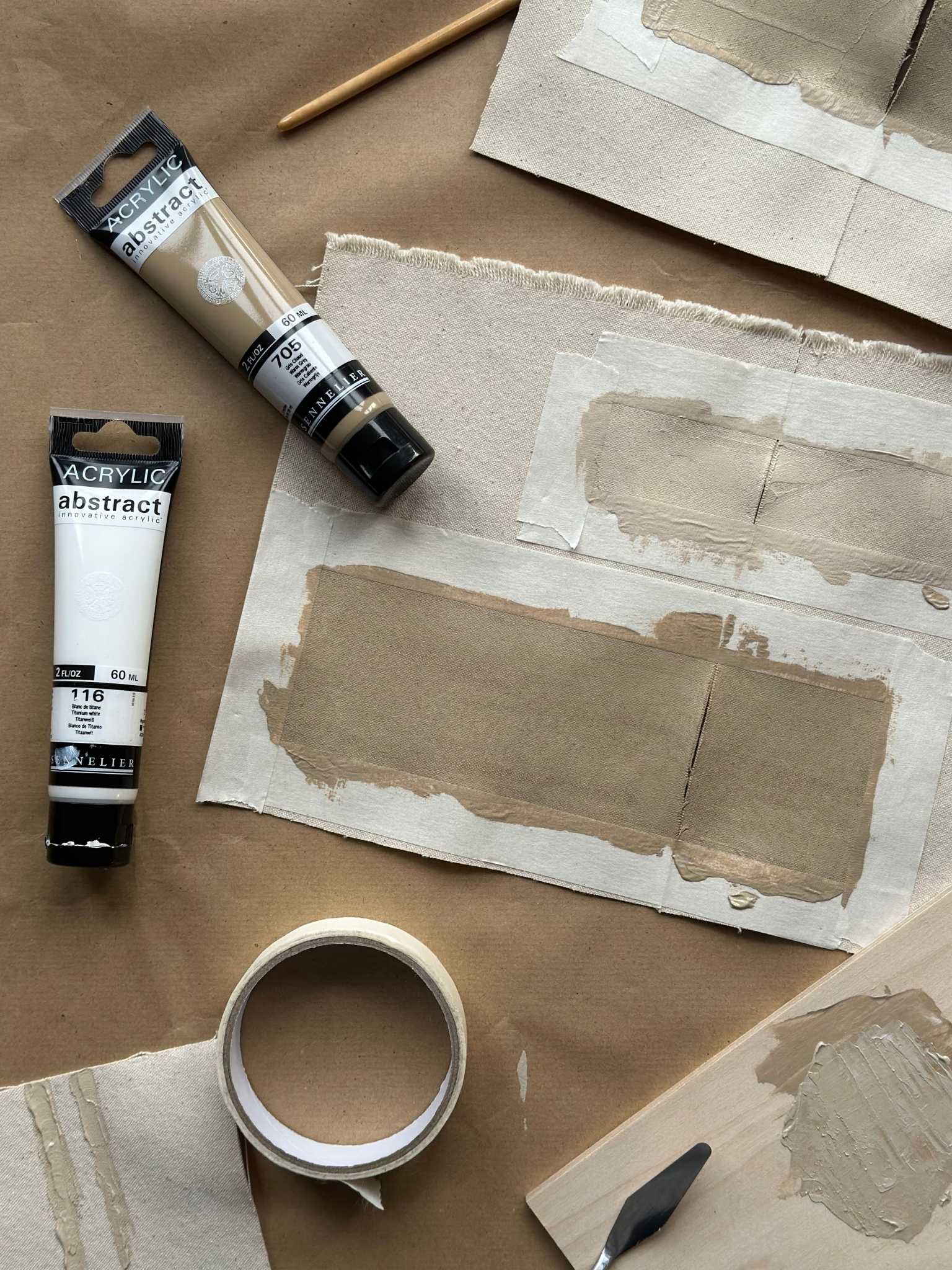 Flat lay process shot showing beige acrylic paint samples applied to cotton canvas test pieces with masking tape, alongside acrylic paint tubes, palette knife and tape.