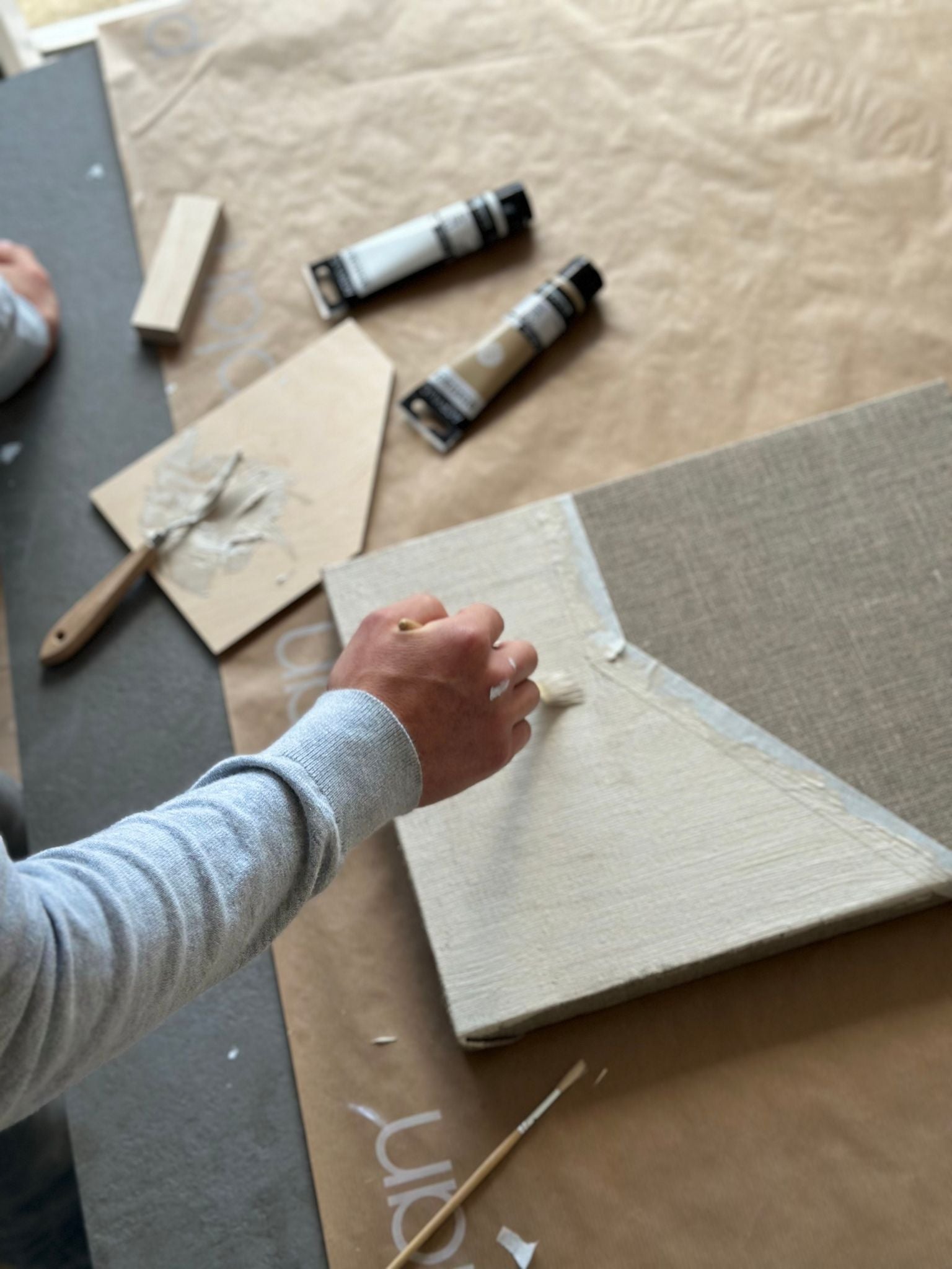Process shot of monochrome linen canvas painting in progress, showing white acrylic paint applied to raw linen canvas using a brush, with paint tubes and tools visible on a protective surface.