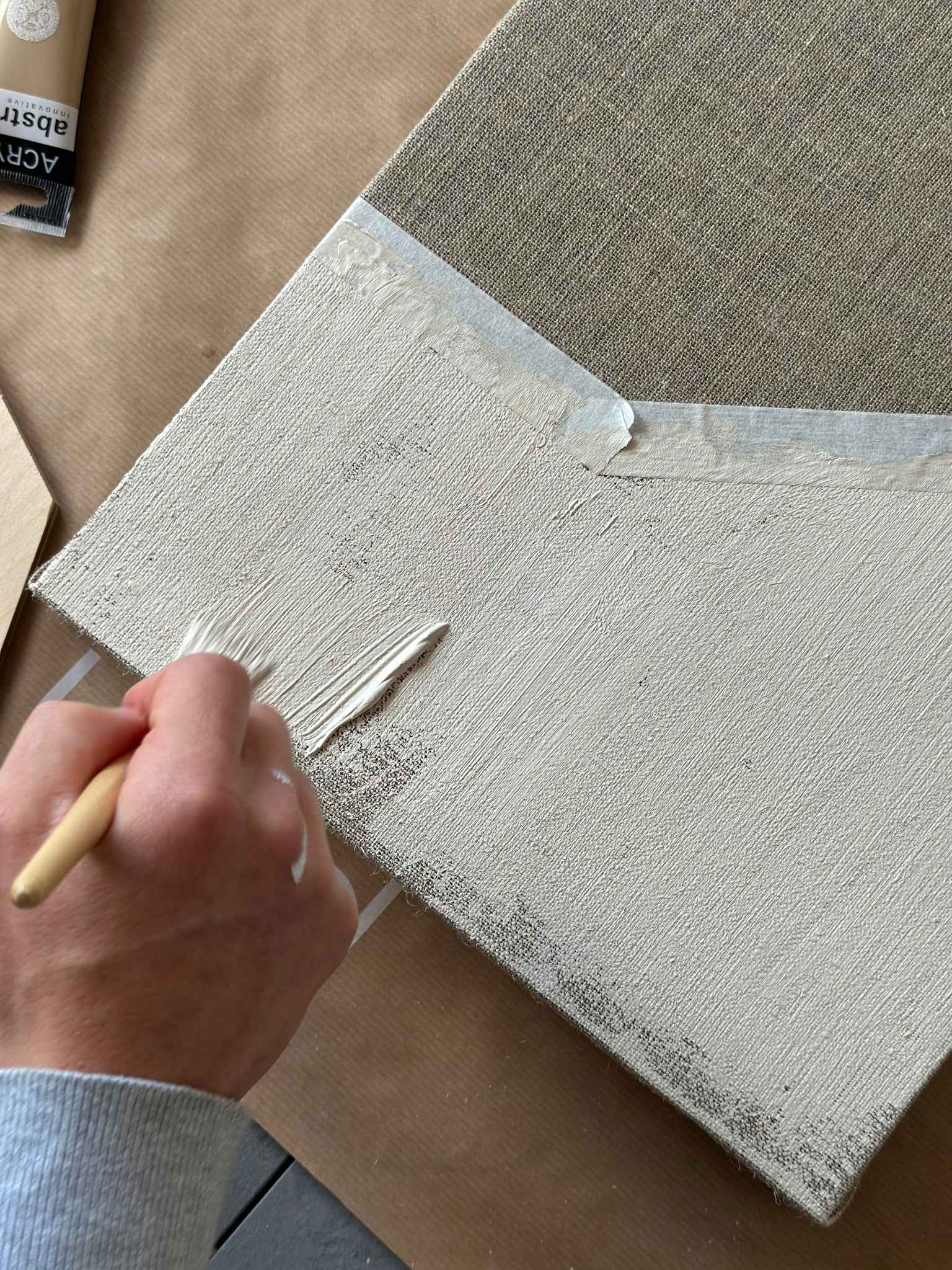 Close-up process shot of beige acrylic paint being brushed onto raw linen canvas, showing textured brushstrokes, masking tape edges and natural linen weave.