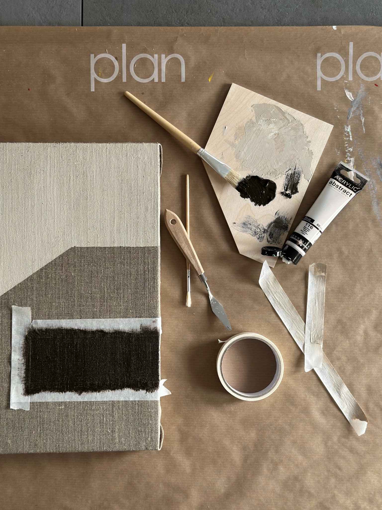 Top-down process shot of monochrome painting setup, featuring a linen canvas with black painted section, palette knives, brushes, masking tape and black and white acrylic paint on a Plan-branded work surface.