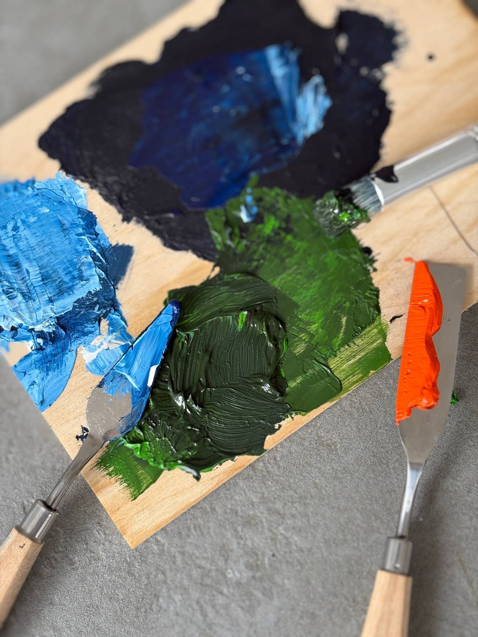 Colour mixing process shot using the Navy & Bright art kit, showing blue, green, black and bright orange acrylic paint on a wooden palette with palette knives.
