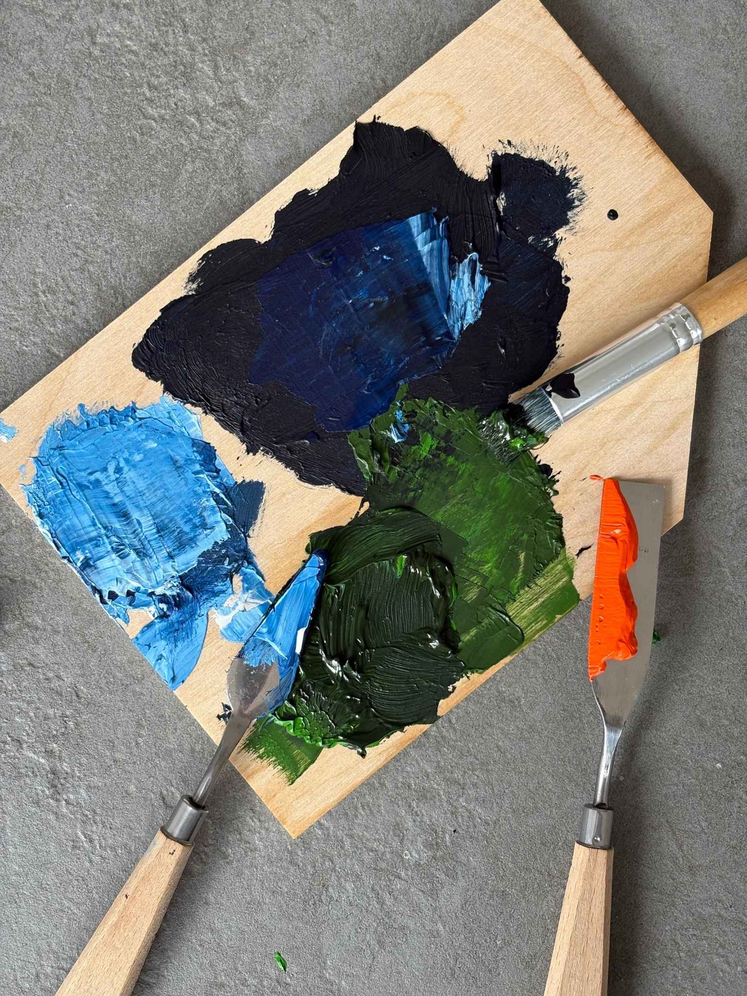 Top-down process shot of mixed navy, blue, green and black acrylic paints on a wooden palette, with palette knives used for colour mixing and preparation.