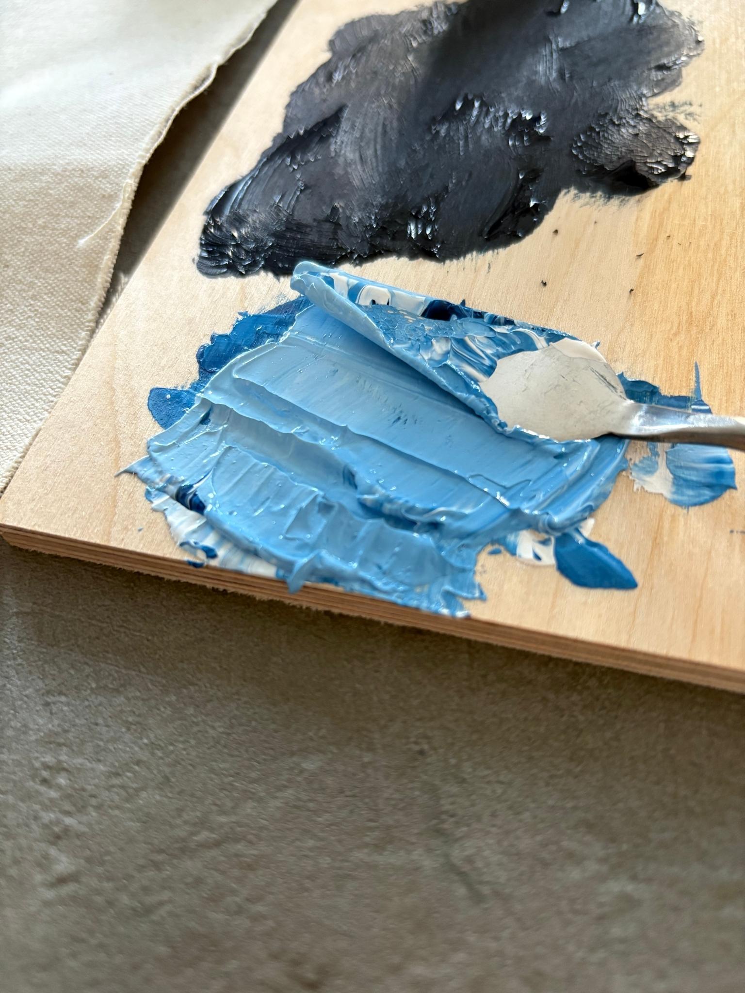 Close-up of light blue acrylic paint being mixed with white on a wooden palette using a palette knife, showing smooth texture and colour blending during the creative process.