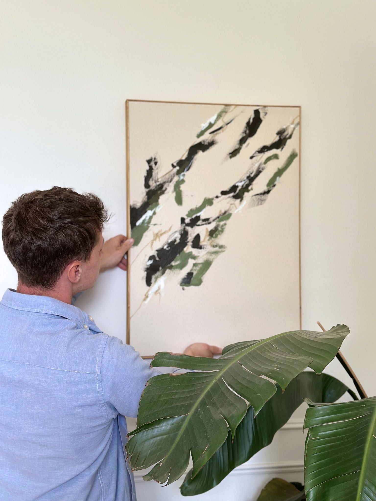 Abstract olive artwork painted on cotton canvas and finished with a handcrafted oak edge frame from the Plan DIY framed canvas art kit, being hung on wall next to indoor plants