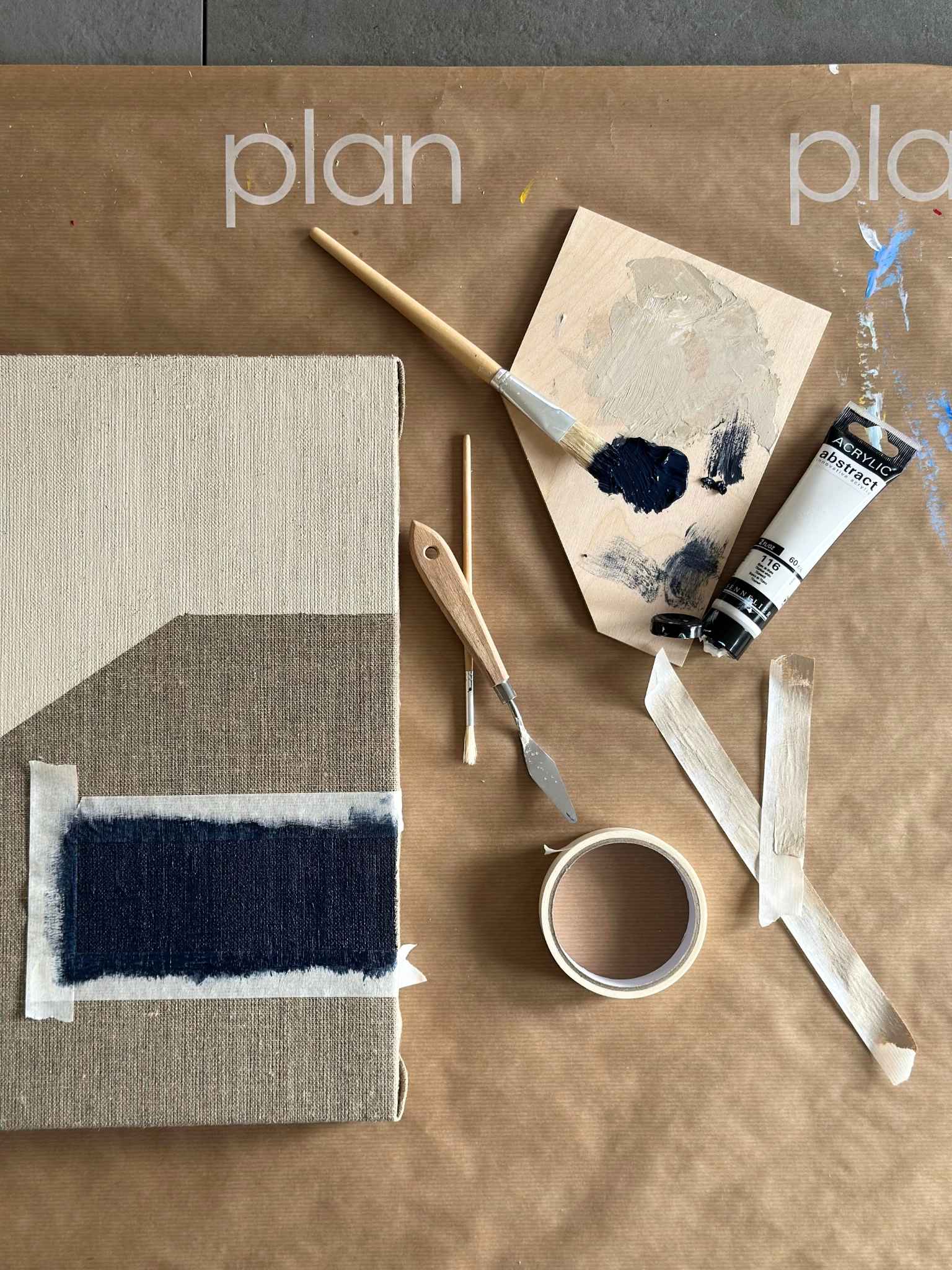 Top-down process shot of monochrome painting setup, featuring a linen canvas with navy painted section, palette knives, brushes, masking tape and black and white acrylic paint on a Plan-branded work surface.