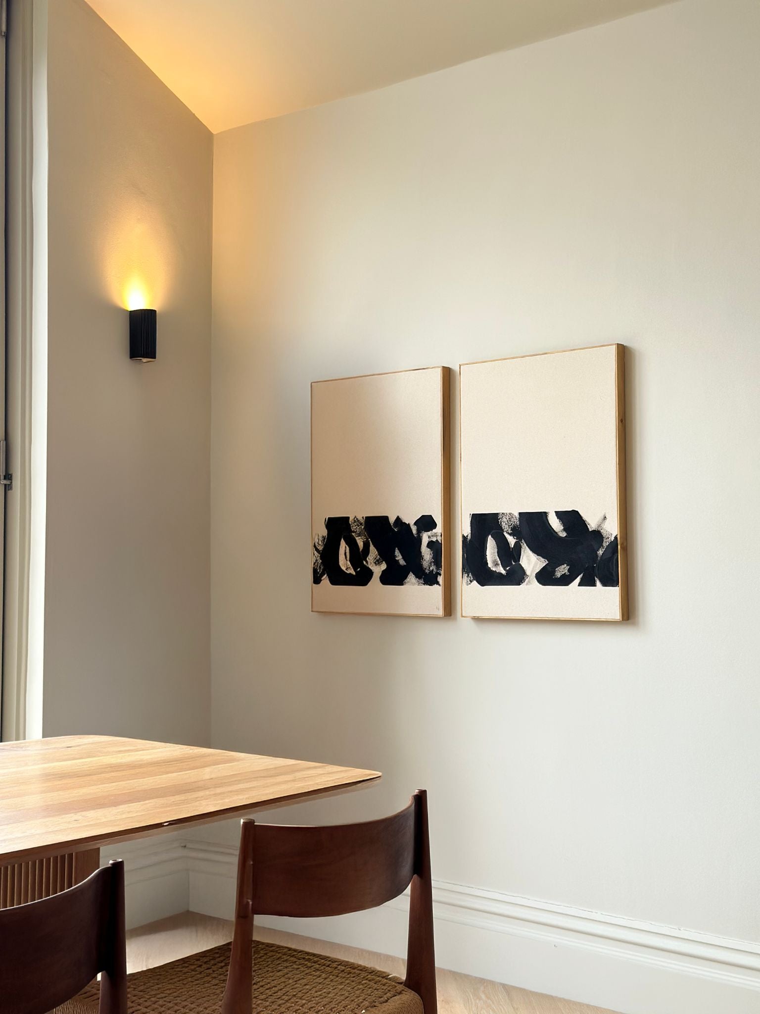 Two framed cotton canvases with bold black monochrome brush marks, hung side by side on a dining room wall above a wooden table with warm wall light.