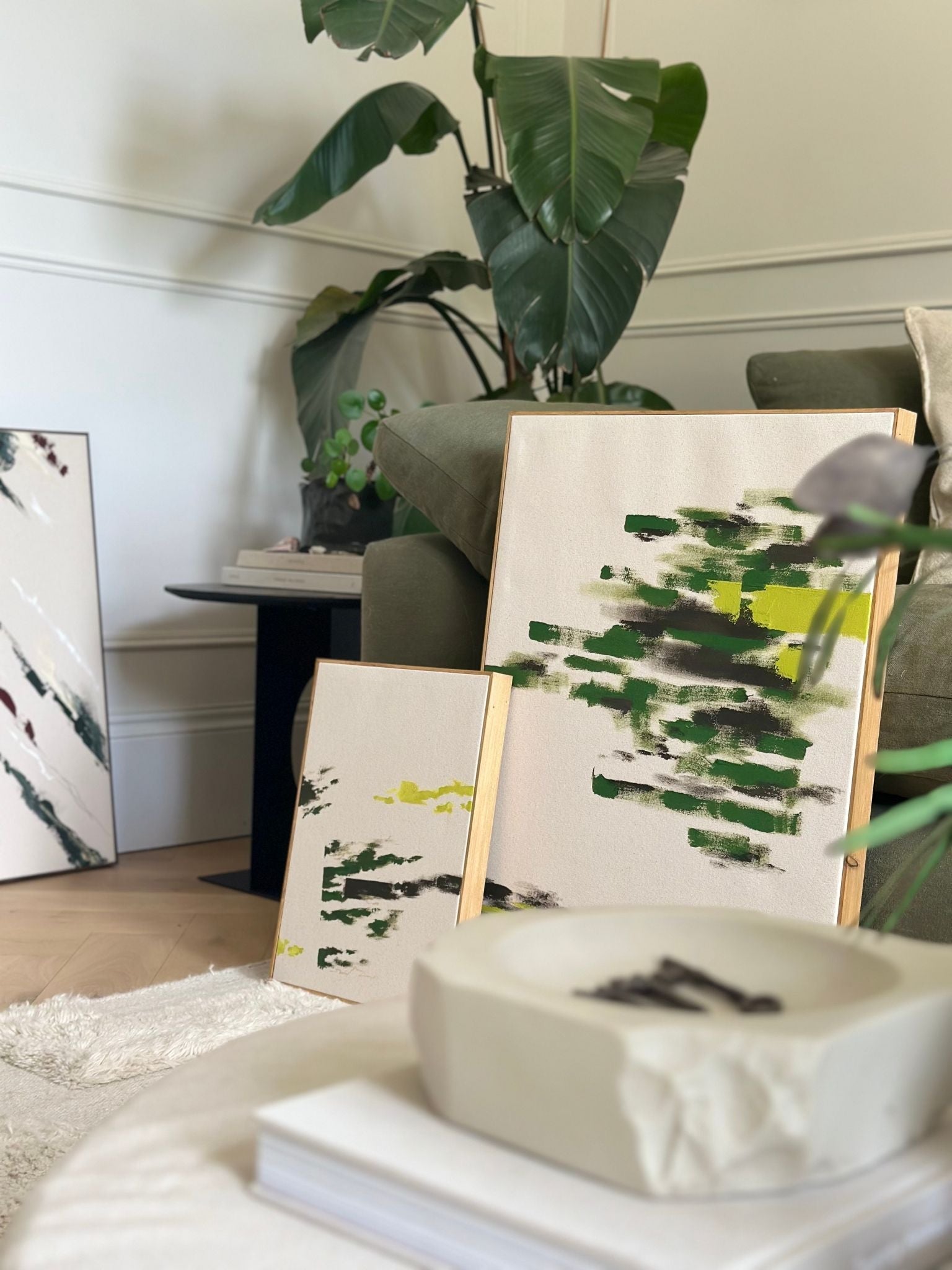 Two framed olive artworks leaning against a wall in a living room setting, featuring expressive green brushstrokes on cotton canvas.