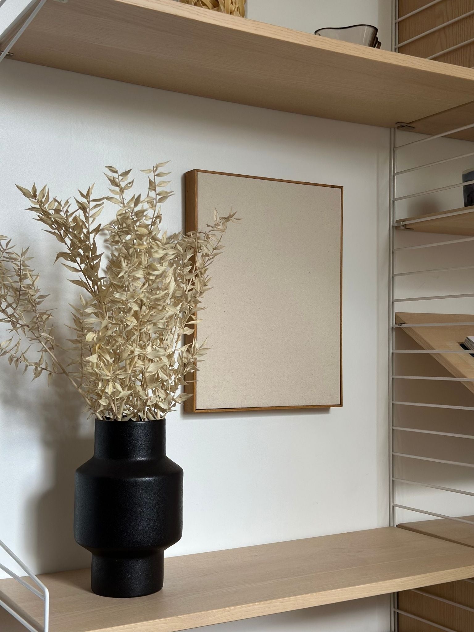 Blank framed cotton canvas artwork hanging on a white wall, styled with shelving and dried stems using a DIY cotton canvas art kit