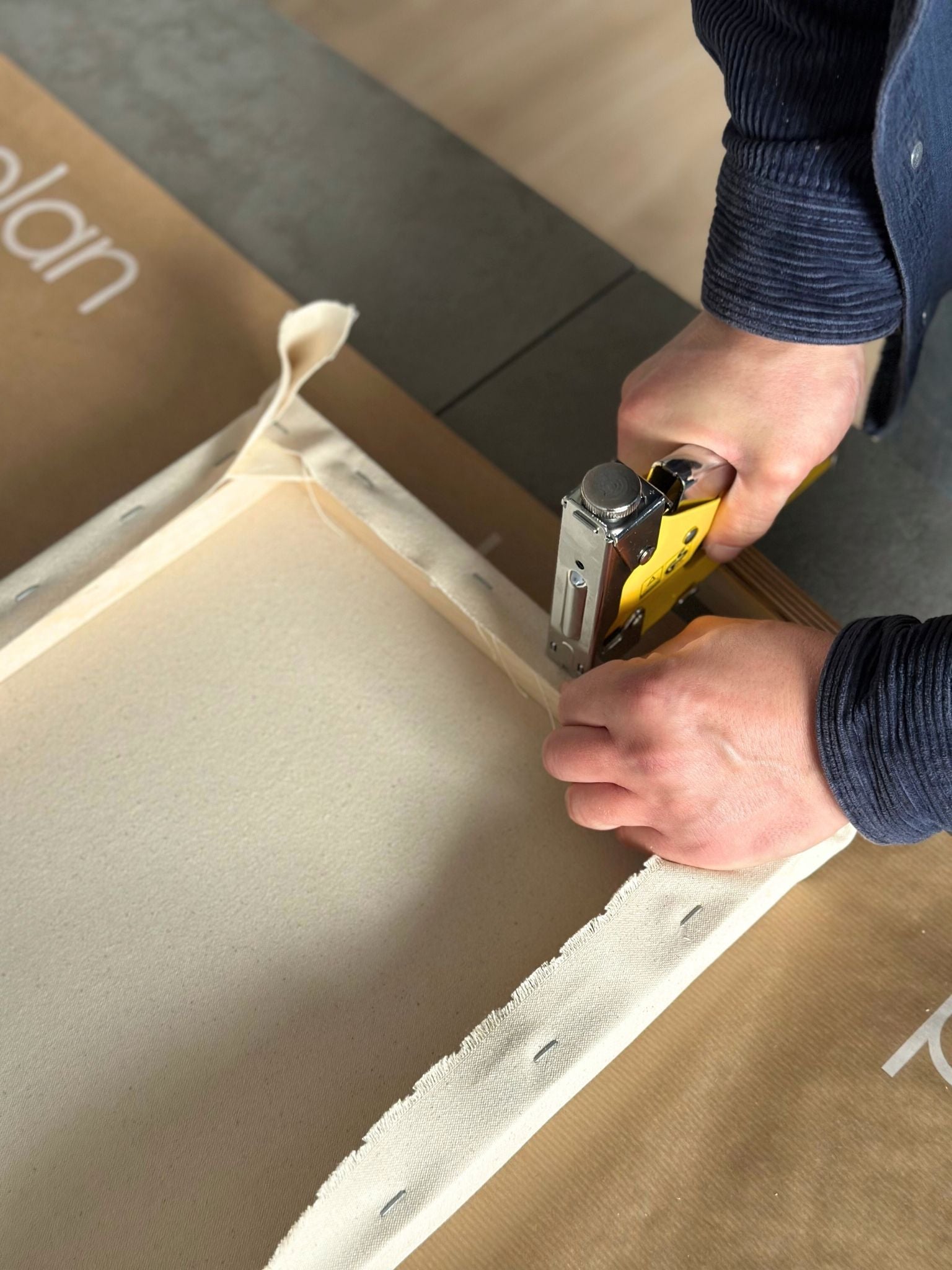 Close-up of cotton canvas being stapled onto a wooden frame using a staple gun as part of a DIY framed canvas art kit