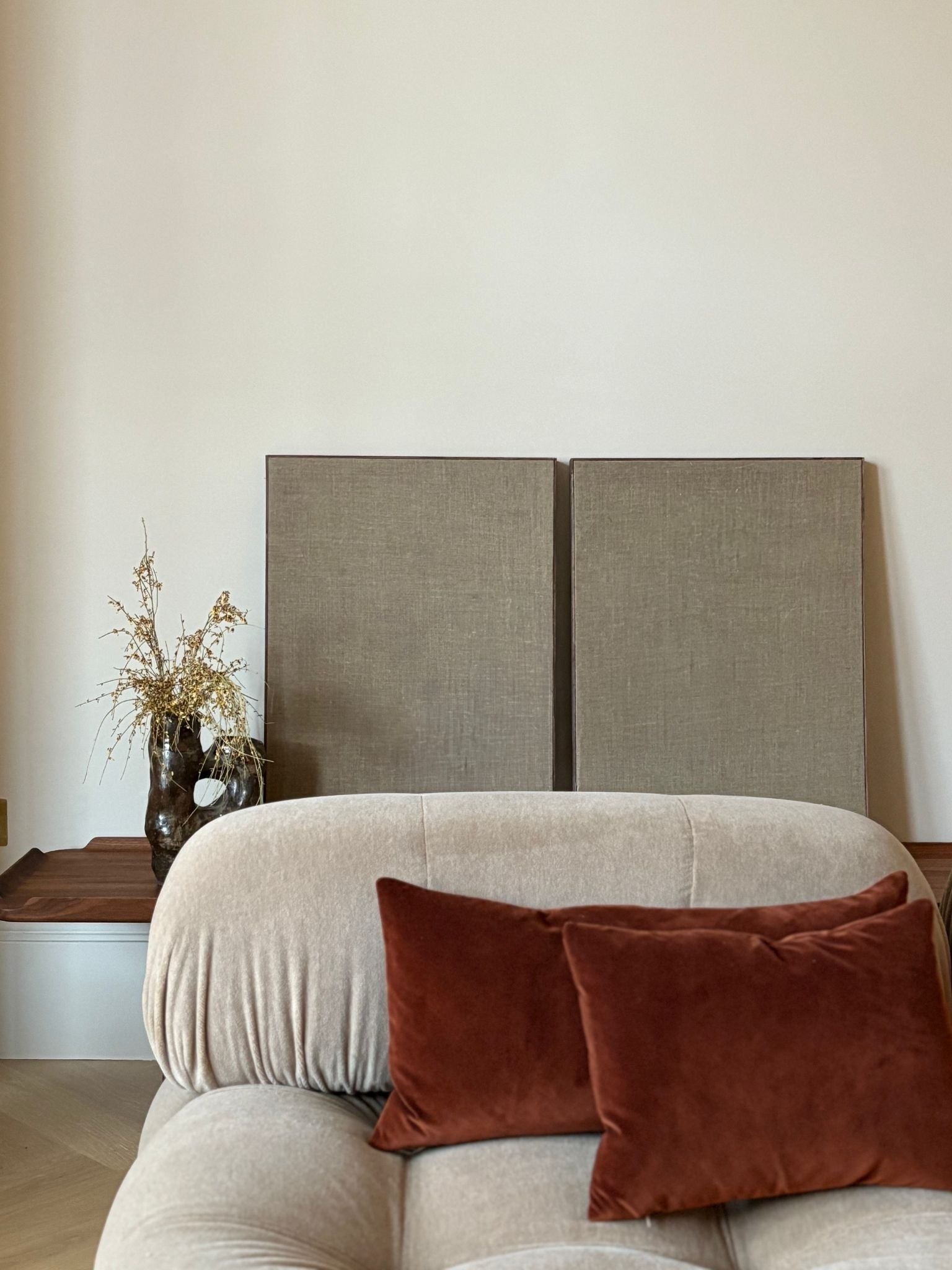 Two blank DIY framed linen canvases leaning on a wooden bench behind a neutral sofa with rust cushions, styled in a minimalist living room.