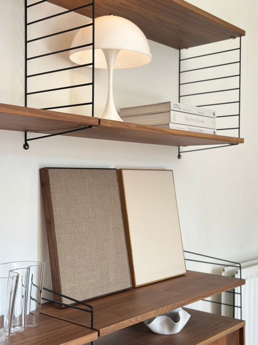 Framed blank canvas artworks resting on wooden shelves, featuring linen and cotton canvases in dark oak and oak edge frames within a modern Scandinavian-style interior.