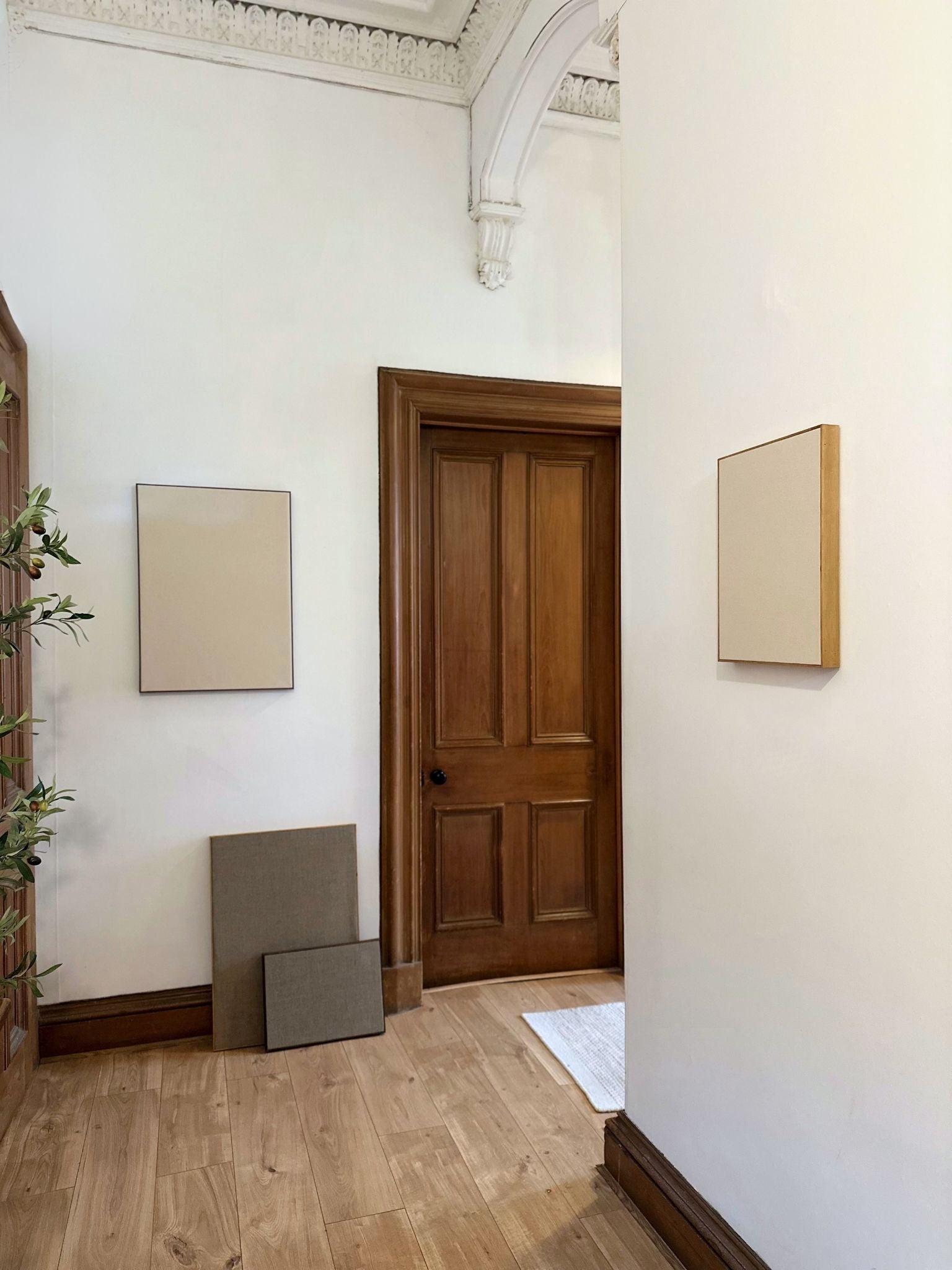 Blank framed canvases styled in a hallway, with one canvas wall-mounted and others leaning on the floor, featuring neutral linen and cotton textures.