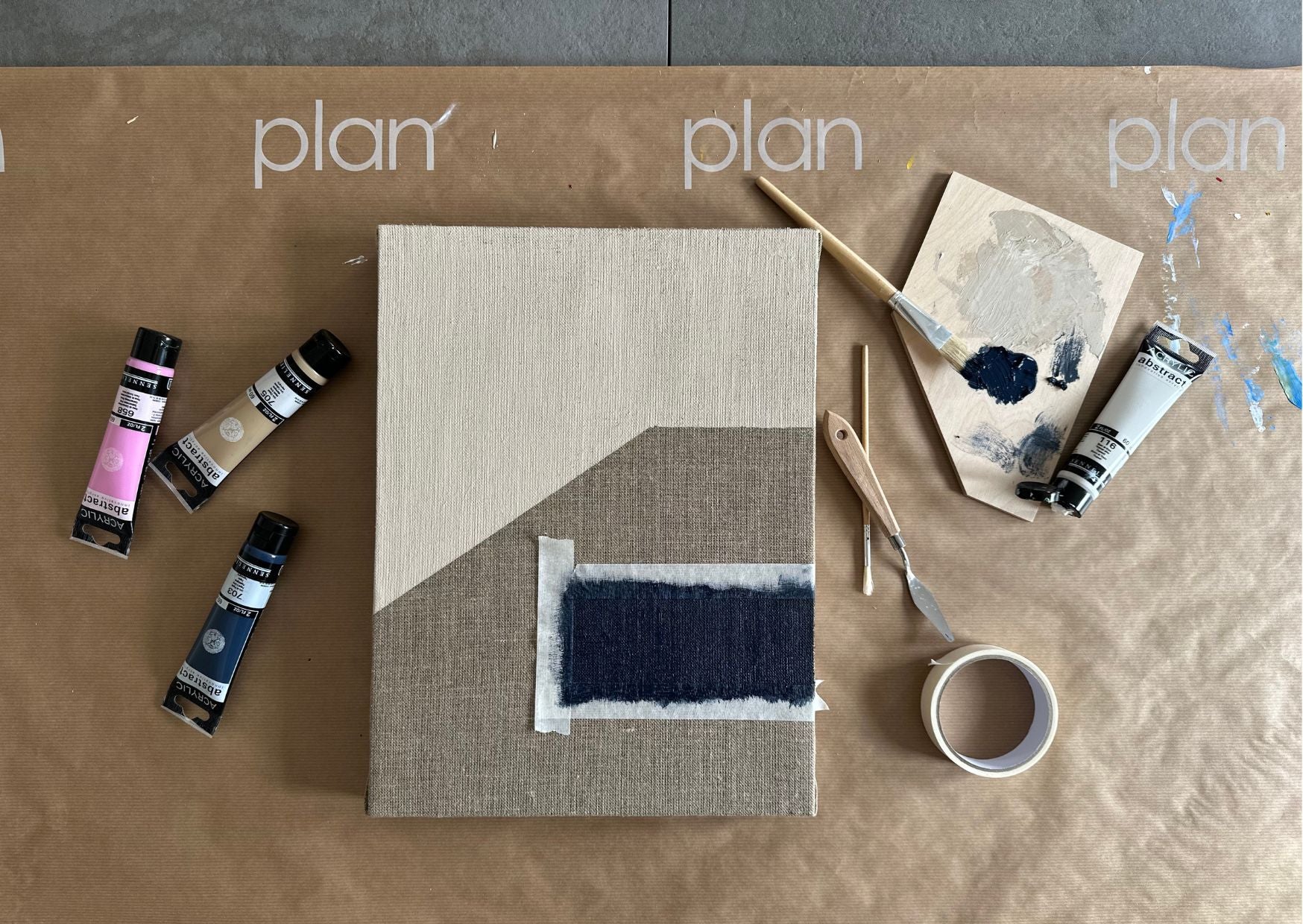 Flat lay of the Plan Process showing a nearly finished abstract artwork on linen canvas, on a Plan-branded surface, surrounded by paint tubes, palette knives, brushes, and masking tape. The image captures the hands-on creative process behind each Plan Art Kit — from building and painting to creating a personal, display-ready artwork.